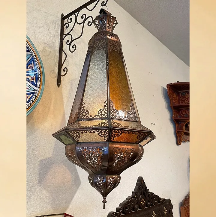 A large, artisanal Moroccan lantern with amber and white glass panels displayed on a wall alongside traditional decor, including a colorful hand-painted ceramic plate and intricately carved wooden wall art.