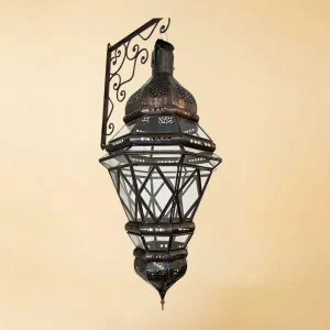 A traditional Moroccan lantern with clear glass panels and bronze-finished metalwork, hanging on a white wall surrounded by other artisanal decor like painted ceramic plates and carved wooden shelves.