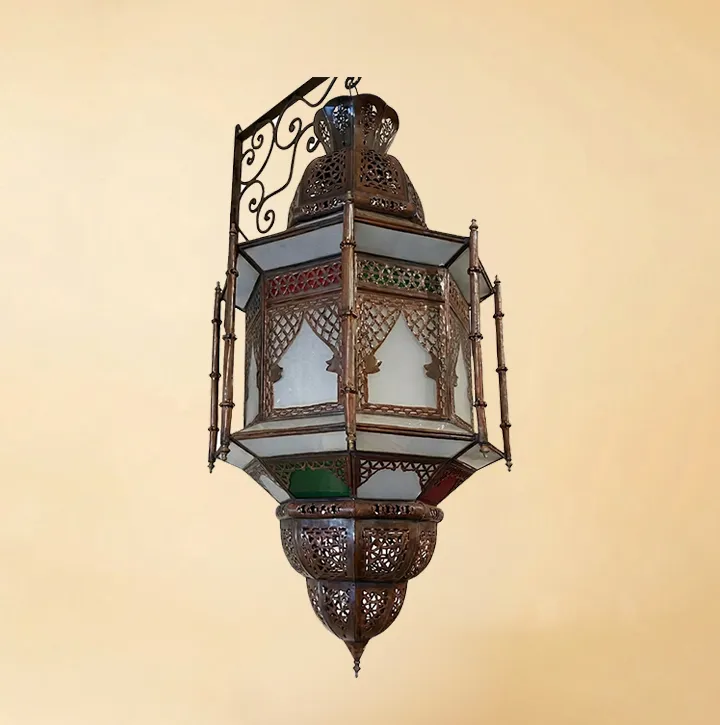 An isolated view of a tall, hexagonal Moroccan lantern featuring white frosted glass panels, small green and red glass accents, and intricate bronze metalwork, suspended from a decorative scrollwork wall bracket.