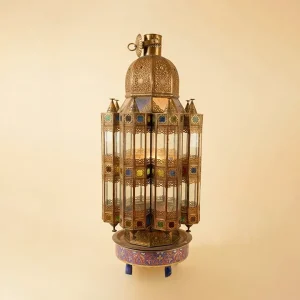 An intricate, tall Moorish-style brass lantern featuring detailed filigree metalwork, multi-colored stained glass inserts, and a domed top, resting on a decorative enameled base against a plain background.