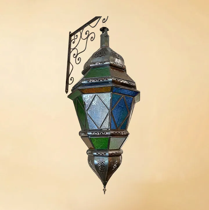 A large, ornate Moroccan hanging lantern with multi-colored stained glass panels in green, blue, and white, featuring intricate bronze metalwork and suspended from a decorative scrollwork wall bracket against a neutral background.