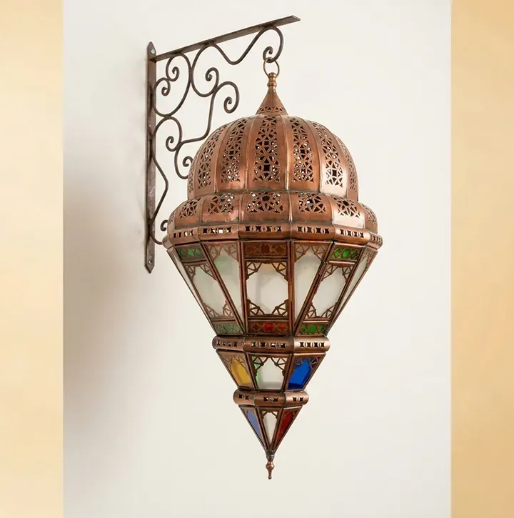 An ornate Moroccan copper lantern with colorful geometric glass panels mounted on a white wall, showcasing the intricate metalwork and traditional artisanal design.